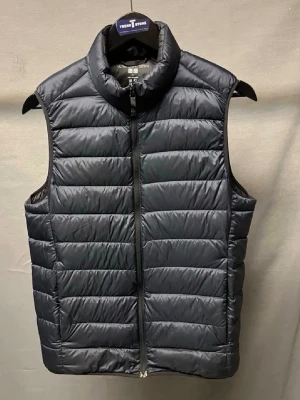 Mörkblå dunväst från Uniqlo - Stilren dunväst från Uniqlo med hög krage. Västen har dragkedja framtill och är lätt vadderad med dun, vilket gör den varm å skön att ha på våren. Perfekt att slänga över en hoodie eller tröja för extra värme. Storlek S