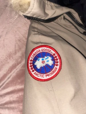 Beige dunjacka från Canada Goose - Säljer en beige dunjacka från Canada Goose med klassisk röd och vit logga på ärmen. Jackan har luva med fuskpäls, svarta detaljer vid fickor och dragkedja samt ribbade muddar. Perfekt för kalla vinterdagar och riktigt snygg streetstil.