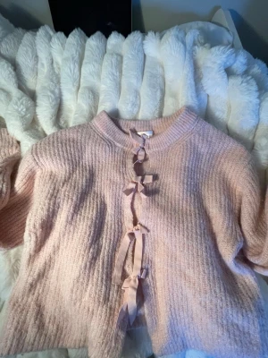 Rosa kofta med rosetter Gina Tricot - Supersöt ljusrosa kofta från Gina Tricot med tre dekorativa rosetter framtill. Koftan är stickad i mjukt material och har rund halsringning. Perfekt för dig som gillar gulliga detaljer och en mysig stil. 