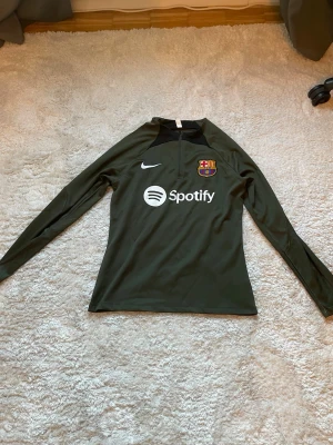 Barcelona träningsset Nike grön - Säljer ett grönt träningsset från Nike och FC Barcelona. Setet består av en långärmad tröja med halv dragkedja och byxor med resår i midjan. Båda plaggen har Barcelona-logga och Nike Swoosh, samt Spotify-logga på tröjan. Perfekt för fotbollsträning eller chill.