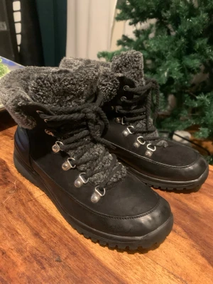 Svarta kängor med fuskpäls och snörning - Säljer ett par svarta kängor med grov sula, snörning och grå fuskpäls runt skaftet. Metallhakar för snörningen ger en cool outdoor-look. Perfekta för kalla dagar och riktigt snygga till jeans eller leggings.