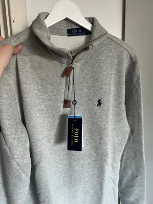 Grå tröja från Polo Ralph Lauren - Snygg ljusgrå tröja från Polo Ralph Lauren med halv dragkedja och ståkrage. Tröjan har det klassiska broderade logotypen på bröstet och ribbade muddar vid ärmslut och nederkant. Perfekt till jeans eller chinos för en clean look.