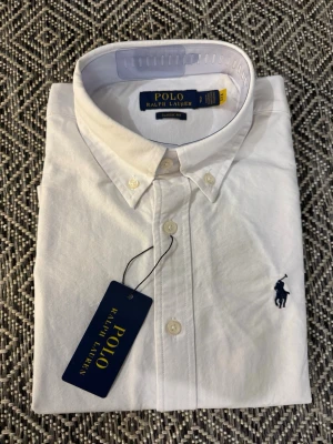 Vit skjorta från Polo Ralph Lauren  - Klassisk vit skjorta från Polo Ralph Lauren i modellen Classic Fit. Skjortan har button-down krage, långa ärmar och den ikoniska mörkblå broderade loggan på bröstet. Tillverkad i mjukt bomullstyg och stängs med vita knappar framtill.