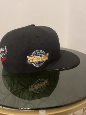 Svart World Series keps - Säljer en svart New Era keps med platt skärm och broderad World Series-logga på sidan. Baksidan har texten 'Love & Hustle = Game' i vitt och rött samt MLB-logga. Klassisk streetwear-stil, perfekt för dig som gillar sport och snygga accessoarer.