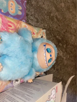 Blå fluffig äkta labubu  - Säljer en söt labubu med blå fluffig päls, stora blå ögon och kaninöron. Ansiktet är beige med ett brett leende och vita tänder. Figurerna är populära som dekoration eller leksak och kommer i originalförpackning med färgglad design.