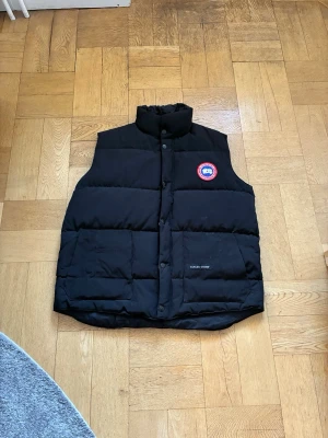 Svart dunväst från Canada Goose - Säljer en svart dunväst från Canada Goose med hög krage och tryckknappar framtill. Västen har två stora fickor och den klassiska röda och vita loggan på bröstet. Perfekt för lager på lager och riktigt varm tack vare dunfyllningen. Skick 9/10