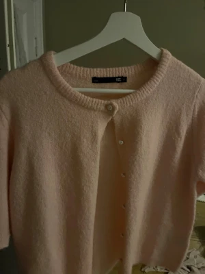 Ljusrosa kofta med rund hals - Mysig ljusrosa kofta från Lager 157 med rund halsringning och knappar framtill. Koftan har en fluffig, mjuk struktur och kortare ärmar. Perfekt för lager på lager och enkel att matcha med olika outfits. Passar både större och mindre storlek på grund av det stretchiga materialet. Använd endast 2 gånger i Spanien. 