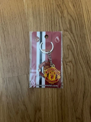 Manchester United nyckelring - Säljer en snygg nyckelring med Manchester Uniteds klassiska klubbmärke i rött, gult och svart. Perfekt accessoar för dig som älskar fotboll och vill visa ditt lag. Metallkedja och ring för enkel fastsättning på nycklar eller väska.