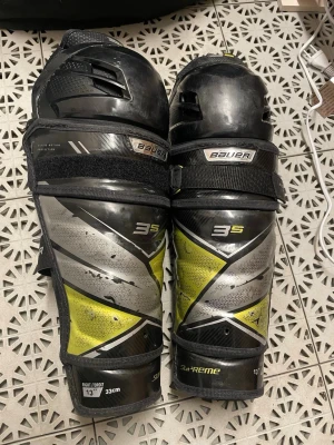 Bauer Supreme 3S benskydd 33 cm - Ett par Bauer Supreme 3S benskydd i svart, silver och gul färg. Skydden har justerbara kardborreband och vadderad insida i gult meshmaterial för extra komfort. Designade för att ge bra skydd och rörelsefrihet vid sportaktiviteter.