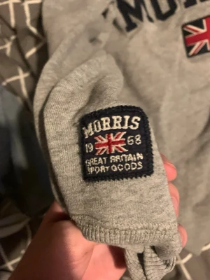 Grå hoodie från Morris med brittisk flagga - Snygg grå hoodie från Morris med broderad brittisk flagga och tydliga loggor på både bröstet och ärmen. Hoodien har klassisk passform, ribbade muddar och är tillverkad i mjukt bomullsmaterial. Perfekt för en avslappnad och stilren look. Pris kan diskuteras skicka ett bud