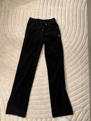 Svarta velourbyxor Juicy Couture - Säljer ett par svarta mjukisbyxor från Juicy Couture i mjuk velour. Byxorna har raka ben, dragsko i midjan och ikoniska Juicy-texten i strass på baksidan. Perfekta för en chill och trendig look.