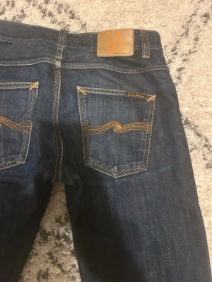 Mörkblå jeans från Nudie Jeans - Säljer ett par mörkblå jeans från Nudie Jeans med klassisk orange söm och vågig detalj på bakfickorna. Jeansen har rak passform och är tillverkade i slitstarkt denimtyg. Perfekta för dig som gillar stilrena och tidlösa jeans.