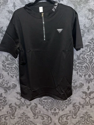 Svart Prada t-shirt med half zip - Snygg svart t-shirt från Prada med kort ärm och half zip framtill. T-shirten har en diskret logga på bröstet och textdetalj i halsen. Tillverkad i ett mjukt material som känns skönt mot huden. Perfekt för dig som gillar stilrena plagg med exklusiv känsla. 