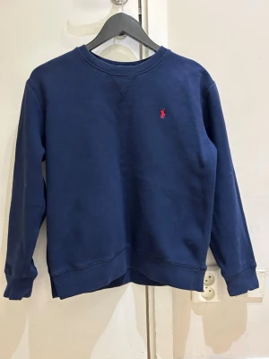 Mörkblå sweatshirt från Ralph Lauren - Mörkblå sweatshirt med rund hals från Ralph Lauren. Klassisk design med liten röd broderad logga på bröstet. Tröjan har långa ärmar och ribbade muddar vid ärmslut och nederkant. Perfekt för en avslappnad och stilren look.
