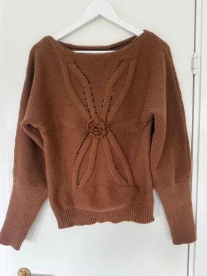 Brun stickad tröja med blomdetalj - Säljer en brun stickad tröja med unik blomapplikation och dekorativa hålmönster framtill. Tröjan har bred halsringning, långa ärmar och ribbade muddar. Perfekt för dig som gillar detaljer och vill ha något extra i garderoben.