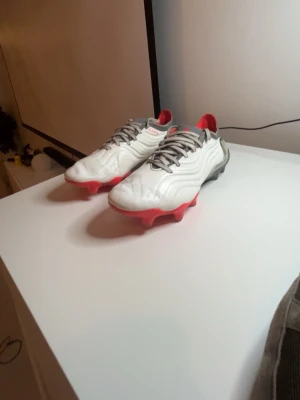 Adidas Copa fotbollsskor vit/röd - Säljer ett par Adidas Copa fotbollsskor med vit ovandel, grå detaljer och röda dobbar. Skorna har texten 'COPA' och 'FUSIONSKIN' på sidan. Designen är modern med vågiga linjer och snörning framtill. Perfekta för dig som vill sticka ut på planen.