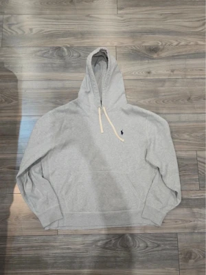 Ralph lauren hoodie - Snygg grå hoodie från Polo Ralph Lauren med klassisk huva och dragsnören i vitt. Broderad blå logga på bröstet och känguruficka framtill. Tillverkad i mjukt bomullsmaterial som är perfekt för en chill och avslappnad stil.