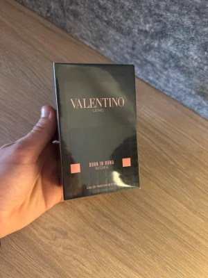 Valentino Uomo Born In Roma Intense - Valentino Uomo Born In Roma Intense Eau de Parfum i en elegant svart rektangulär förpackning med guldiga och rosa detaljer. Flaskan rymmer 100 ml och har en lyxig och modern design som passar perfekt för dig som gillar exklusiva dofter.
