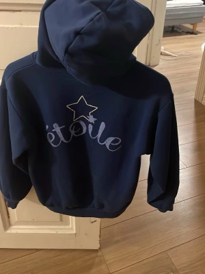 Mörkblå hoodie med stjärntryck - Snygg mörkblå hoodie med huva och trycket 'étoile' samt en stjärna på ryggen. Mjuk och skön modell med ribbade muddar vid ärmslut och midja. Perfekt för dig som gillar en stilren men ändå cool look.