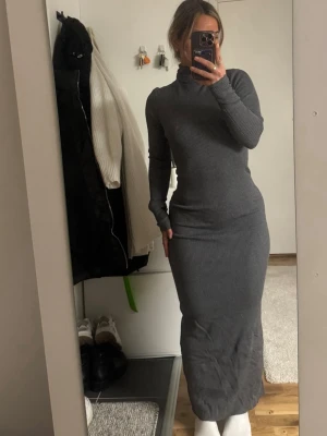 Grå ribbad långklänning med slits - Säljer en grå ribbad långklänning med polokrage och lång ärm. Klänningen har en tight passform och en snygg slits baktill. Perfekt för dig som gillar stilrena och bekväma plagg. Endast testad!