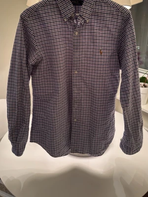 Rutig skjorta från Polo Ralph Lauren - Klassisk långärmad skjorta från Polo Ralph Lauren i blåvitt rutigt mönster. Skjortan har knappar framtill, button-down krage och den ikoniska broderade loggan på bröstet. Perfekt för en stilren och avslappnad look.