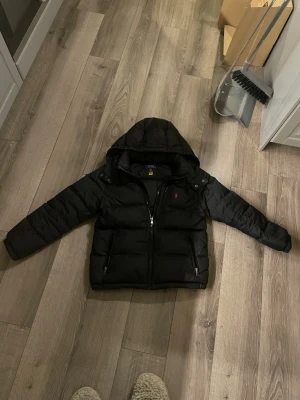 Svart pufferjacka från Polo Ralph Lauren - Säljer en svart pufferjacka från Polo Ralph Lauren med huva och dragkedja framtill. Jackan har två framfickor med dragkedja och den klassiska röda Polo-loggan på bröstet. Perfekt för kalla vinterdagar och riktigt skön att bära.