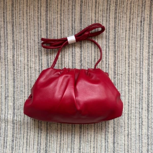 Röd clutchväska - Trendig röd handväska i mjukt, slätt veganskt skinn med puffig form och dragskoöppning. Väskan har en lång, avtagbar axelrem som kan bäras crossbody eller på axeln. Perfekt för dig som vill ha en statementväska med modern look.