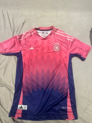 Adidas rosa/blå Tyskland matchtröja - Snygg matchtröja från Adidas i rosa och blått med zigzag-mönster. Tysklands landslagsmärke och fyra stjärnor på bröstet, samt HEAT.RDY- och Aeroready-teknologi för bra ventilation. Korta ärmar och sportig passform, perfekt för fotboll eller träning.