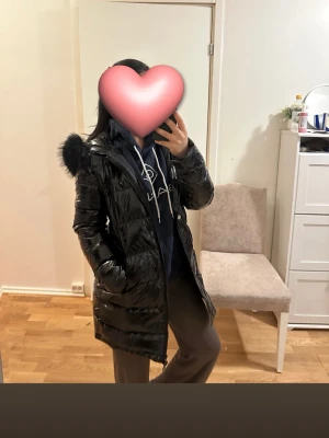 Svart glansig jacka med pälshuva - Säljer en svart, glansig dunjacka med huva och pälskant. Jackan är lång, har dragkedja framtill och två stora fickor på sidorna. Perfekt för kalla vinterdagar och riktigt snygg streetstil,andvänd ett par gånger bara så är exakt som ny,pris kan diskuteras 
