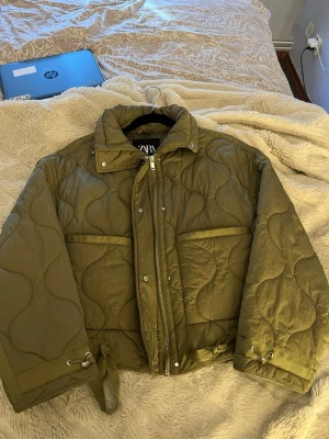 Quiltad jacka från Zara - Säljer en olivgrön pufferjacka från Zara med vågigt quiltat mönster, hög krage och dragkedja framtill. Jackan har två stora fickor och justerbara band vid ärmsluten för extra stilpoäng. Perfekt för kalla dagar när du vill vara både varm och trendig.