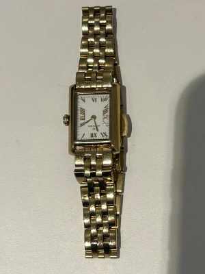 Guldfärgad klocka från Mockberg - Stilren guldfärgad klocka från Mockberg med rektangulärt vitt ur och romerska siffror. Länken är i metall med polerad finish och klassisk design. Perfekt accessoar för dig som gillar tidlös och elegant stil.