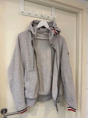 Grå Moncler hoodie med dragkedja - Snygg grå hoodie från Moncler med dragkedja, huva med dragsko och Moncler-logga på ärmen. Ribbade muddar med röd, vit och svart rand. Praktiska fickor framtill och mjukt material som känns skönt mot huden. Perfekt för en avslappnad och trendig look.