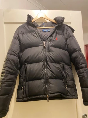 Svart pufferjacka från Polo Ralph Lauren - Snygg svart pufferjacka från Polo Ralph Lauren med huva, dragkedja framtill och två stora fickor med dragkedja. Jackan har en röd broderad logga på bröstet och är perfekt för kalla vinterdagar. Klassisk och stilren design som håller dig varm.
