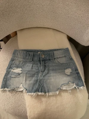 Ljusblå jeanskjol med slitningar - Trendig ljusblå jeanskjol i kort modell med slitna detaljer och fransig nederkant. Kjolen har klassiska fickor fram och bak. Passar som xs/xxs