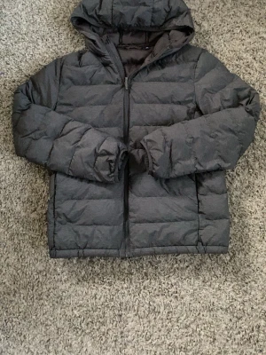 Mörkgrå pufferjacka från Uniqlo - Säljer en mörkgrå pufferjacka från Uniqlo med huva och dragkedja framtill. Jackan har en klassisk quiltad design och är tillverkad i ett lätt, vadderat material som håller värmen. Perfekt för kalla dagar och enkel att matcha med olika outfits.