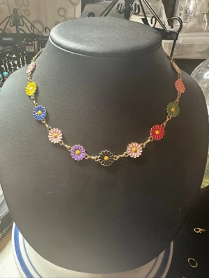 Färgglatt blomhalsband i metall - Superfint halsband med små blommor i olika färger som gult, blått, rosa, lila, svart, grönt och rött. Blommorna sitter på en tunn guldfärgad kedja. Perfekt för dig som gillar färg och vill sticka ut med din stil.