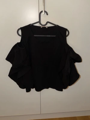 Svart volangtopp från BSB Jeans - Svart topp från BSB Jeans med stora volanger på ärmarna och offshoulder-design. Toppen har en rund halsringning och är tillverkad i ett mjukt tyg som känns bekvämt mot huden. Perfekt för dig som vill sticka ut med en cool och trendig look.