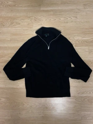 Svart halvzip tröja med hög krage - Stilren svart tröja med halvzip och hög krage. Långa ärmar och ribbade muddar ger en clean look. Perfekt för lager på lager och funkar till det mesta. Materialet ser ut att vara mjukt och bekvämt, troligen bomull eller bomullsblandning.