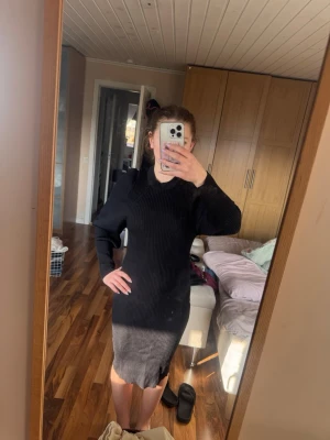 Svart ribbad stickad klänning - Svart stickad klänning med ribbad struktur och långärmad design. Klänningen har en slits bak och en kroppsnära passform som framhäver siluetten. Perfekt för dig som gillar en enkel och stilren look.