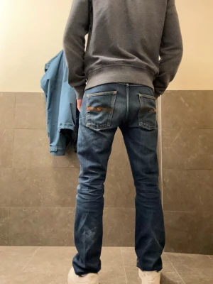Mörkblå jeans från Nudie Jeans - Säljer ett par mörkblå jeans från Nudie Jeans med klassisk femficksdesign och snygga slitningar. Jeansen har raka ben och kontrastsömmar på bakfickorna. Perfekta för en avslappnad och stilren look. Littäe lagat nära bakfickan men det syns knappt