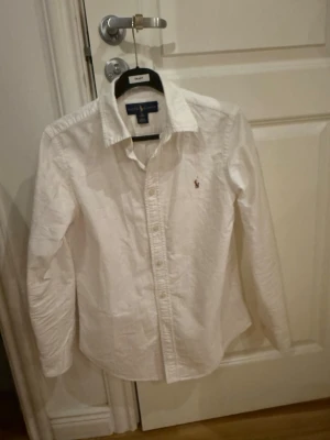 Vit skjorta från Ralph Lauren - Klassisk vit skjorta från Ralph Lauren med broderad logga på bröstet. Skjortan har lång ärm, knappar framtill och en snygg krage. Custom fit-modell i mjuk bomull, perfekt för en stilren look.