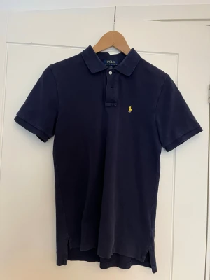 Mörkblå pikétröja från Ralph Lauren - Klassisk mörkblå pikétröja från Ralph Lauren med gul broderad logga på bröstet. Tröjan har korta ärmar, krage och knappar framtill. Tillverkad i mjuk bomull som känns skön mot huden. Perfekt till jeans eller shorts för en clean look. Storlek 14-16 år