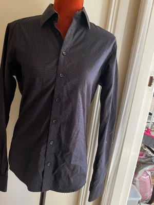 Dressmann svart skjorta, storlek S - Svart långärmad skjorta med ett diskret prickigt mönster. Skjortan har en klassisk krage och knäppning framtill. Ärmsluten är försedda med knappar. Insidan av kragen har ett kontrasterande mönster i svart och vitt. Bomull Slim fit längd: 70 cm Bystvidd: 48 cm  Inga bilder när den sitter på mig! Katter finns i hemmet!