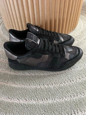 Valentino svarta camo sneakers - Snygga svarta sneakers från Valentino med camo-mönster i olika nyanser av svart och grått. Skorna har detaljer i mocka och läder, svarta skosnören och en cool metallisk häl. Perfekta för dig som vill ha en unik och trendig look.