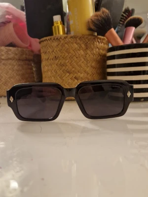 Chrome Hearts glasögn - Snygga svarta solglasögon från Chrome Hearts med rektangulära bågar och mörka glas. Sidorna har silverfärgade detaljer med liljemotiv som ger en unik touch. Perfekta för dig som vill sticka ut med accessoarerna. Perfekta inför våren o sommarn