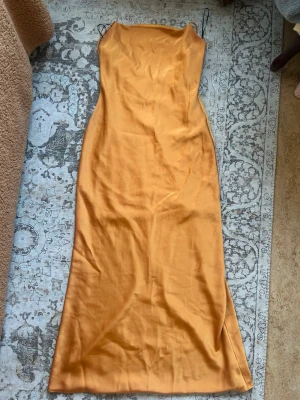 Lång orange slipklänning med smala band - Säljer en lång, figurnära slipklänning i orange med smala axelband och rak urringning. Klänningen har en minimalistisk design och är tillverkad i ett glansigt material som ger en lyxig känsla. Perfekt för sommarens alla tillfällen!