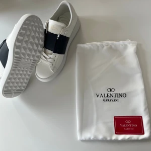 Valentino Garavani vita sneakers - Stilrena vita sneakers från Valentino Garavani med bred svart läderdetalj över snörningen. Skorna har klassisk låg siluett, vita snören och diskret logga på plösen. Yttermaterialet är slätt läder och sulan har ett rutmönstrat grepp.