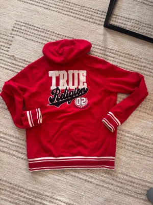 Röd hoodie från True Religion - Snygg röd hoodie från True Religion med broderad logga på bröstet och stor text på ryggen. Tröjan har vita och svarta ränder på muddar och nederkant samt klassisk huva med dragsko. Perfekt för en streetwear-look. I storlek XL, men satt bra på mig som har vanligtvis storlek S, skulle säga att den är mer som M storlek 