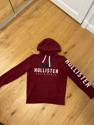 Vinröd hoodie från Hollister - Snygg vinröd hoodie från Hollister med stor logga och text på bröstet samt ärmen. Klassisk känguruficka framtill och vita dragsnören i huvan. Tillverkad i mjukt material som är skönt att ha på sig. Perfekt för en avslappnad och trendig look.