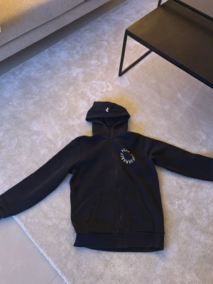 Svart hoodie från Peak Performance - Snygg svart hoodie från Peak Performance med dragkedja framtill och två stora fickor. Märkets logga sitter på bröstet i vitt och även en liten logga på huvan. Perfekt för en avslappnad och sportig stil.